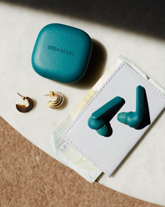 Blue colored Urbanears wireless earbuds sitting on a wallet next to two small gold earrings on a table, representing listening to a podcast