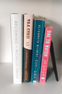 Tall Stack of books containaing Becoming by Michelle Obama, One to Watch by Kate Stayman-London, City of Girls by Elizabeth Gilbert, Transcendent Kingdom by Yaa Gyasi, and an Amazon Nook e-reader