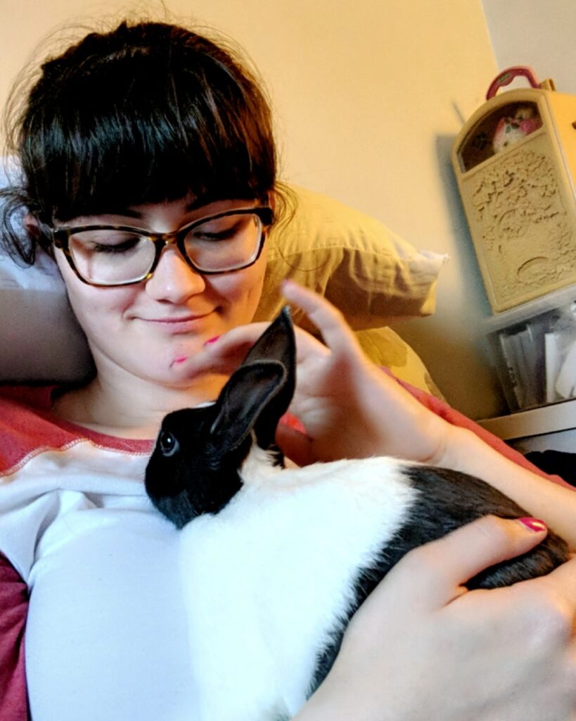 Young Moira, girl with banks and glasses holding baby Black and white Dutch bunny rabbit who inspired the Rabbit Care Guide in her arms and petting his nose.