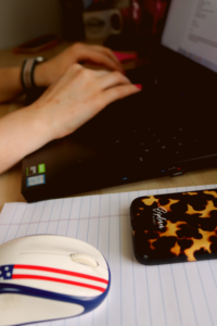 Hands typing on a Dell laptop in the background. In the foreground a white computer mouse with an American flag pattern sits on a white legal pad next to a tortoise shell phone case with Explorer Media printed on it. 