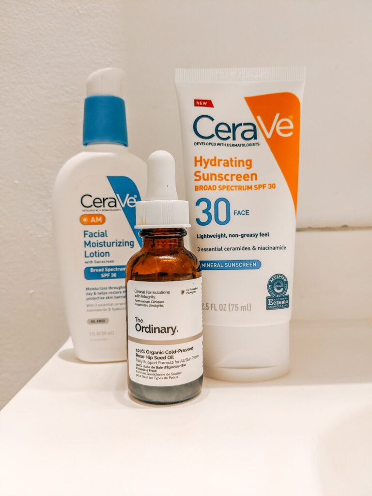 Three budget-friendly summer skincare products: Blue and white bottle of CeraVe AM Facial Moisturizing Lotion, orange and white bottle of CeraVe Hydrating Mineral Sunscreen, and The Ordinary Cold-Pressed Rose Hip Seed Oil.