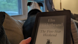 Woman's hand holding a Kindle ereader with the cover of The Five Star Weekend by Elin Hilderbrand while a dog sits on a couch in the background looking out the window