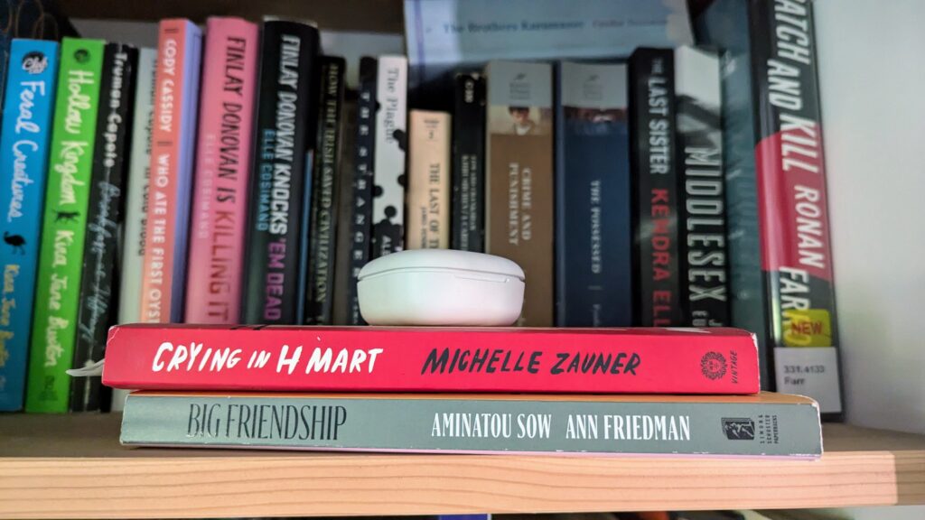 Books Crying in H Mart and Big Friendship stacked on a bookshelf with an ear bud/airpod case on top with other blurred book spines visible in the background